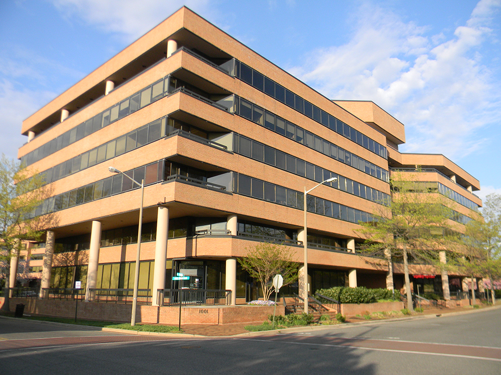 A modern, multi-story office building with tan brick exterior, large horizontal windows, and columns at the entrance, surrounded by trees and a clear blue sky. A street and sidewalk are in the foreground.