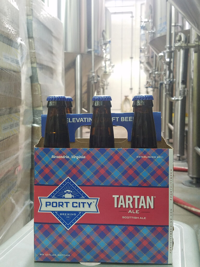 A six-pack of Port City Tartan Ale with blue caps sits on a cart in a brewery, surrounded by large stainless steel tanks. The blue and red plaid packaging prominently displays the brewery’s name and ale variety.