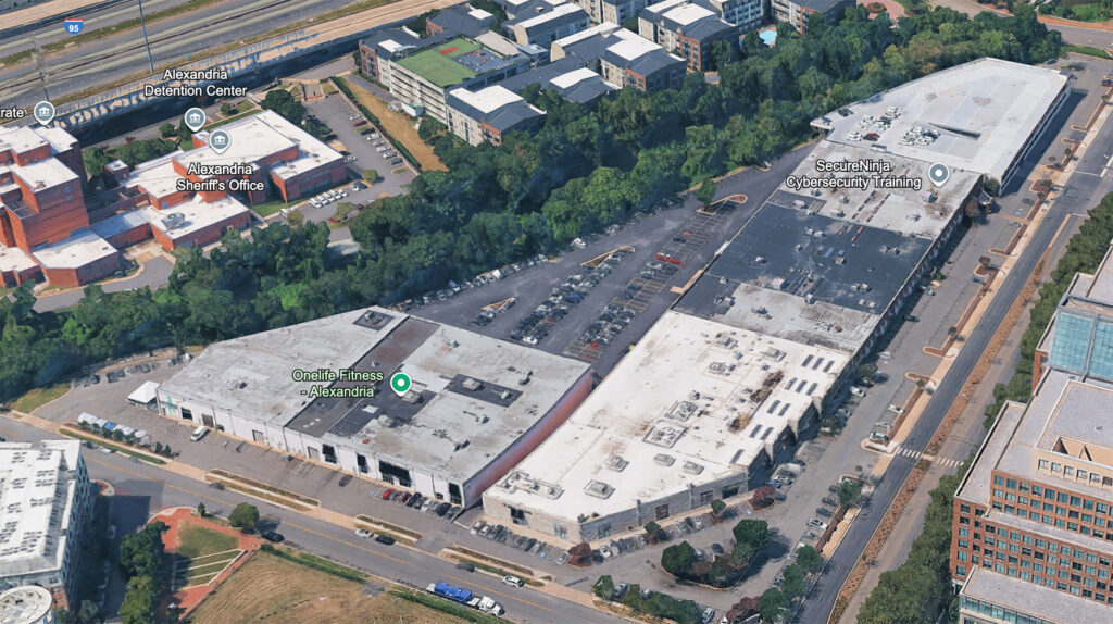 Aerial view of a commercial area with large white-roofed buildings, a parking lot with cars, trees surrounding the lot, and nearby labeled locations including a gym, sheriffs office, and a cybersecurity training center.