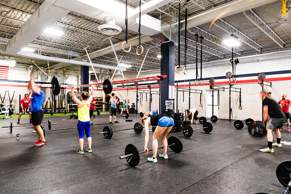 People are lifting barbells and exercising in a spacious, brightly lit gym with high ceilings, gymnastic rings, and various workout equipment. Multiple individuals perform lifts, while others watch or rest.