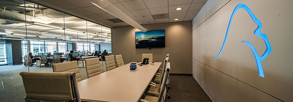 Modern conference room with beige chairs around a long white table, a blue lion outline logo on the wall, glass walls revealing a spacious office area, and a large monitor displaying a scenic image.
