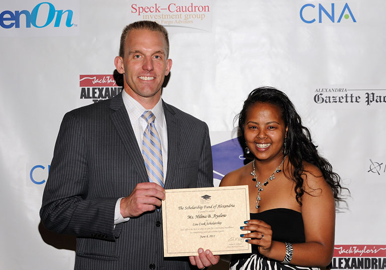 A man in a suit and a young woman in a black dress smile at the camera as they hold a certificate together, standing before a Tartan Properties sponsor backdrop.
