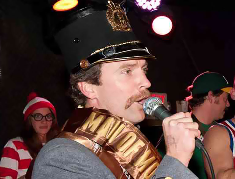 A man with a mustache, wearing a tall official hat and a Tartan Properties sash, speaks into a microphone. In the background, a woman dressed as Waldo from Wheres Waldo? stands among other costumed people.