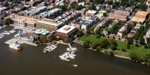 Aerial view of a waterfront town with a marina, multiple docks, boats, green park space, and buildings arranged in blocks near the shoreline. Trees and streets are visible throughout the urban area.