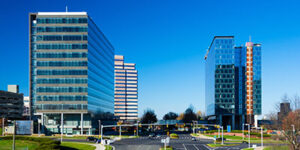 Modern office buildings with glass facades surround a mostly empty parking lot under a clear blue sky. Trees and landscaped grounds are visible between the buildings.