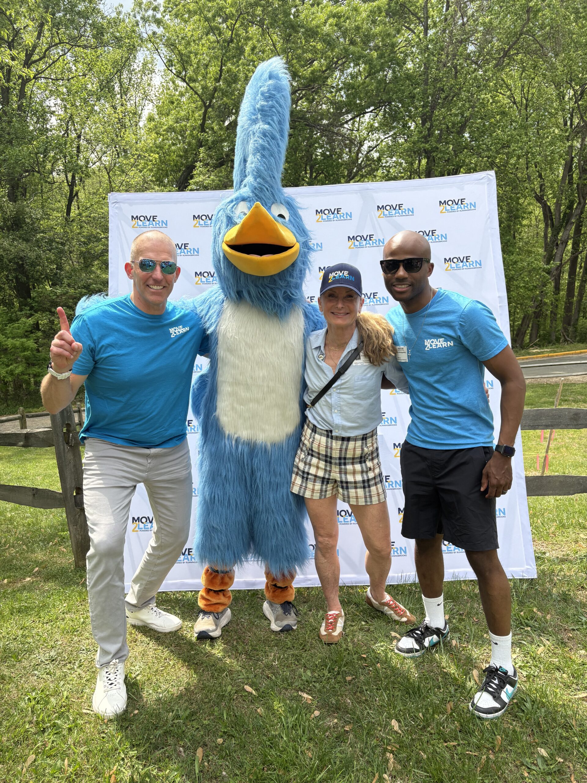 Three adults in blue Move4MHL shirts, alongside a person in a tall, blue bird mascot costume, pose outdoors on green grass with trees behind them. The sunny scene features a Move4MHL and Tartan Properties backdrop as they all smile brightly.