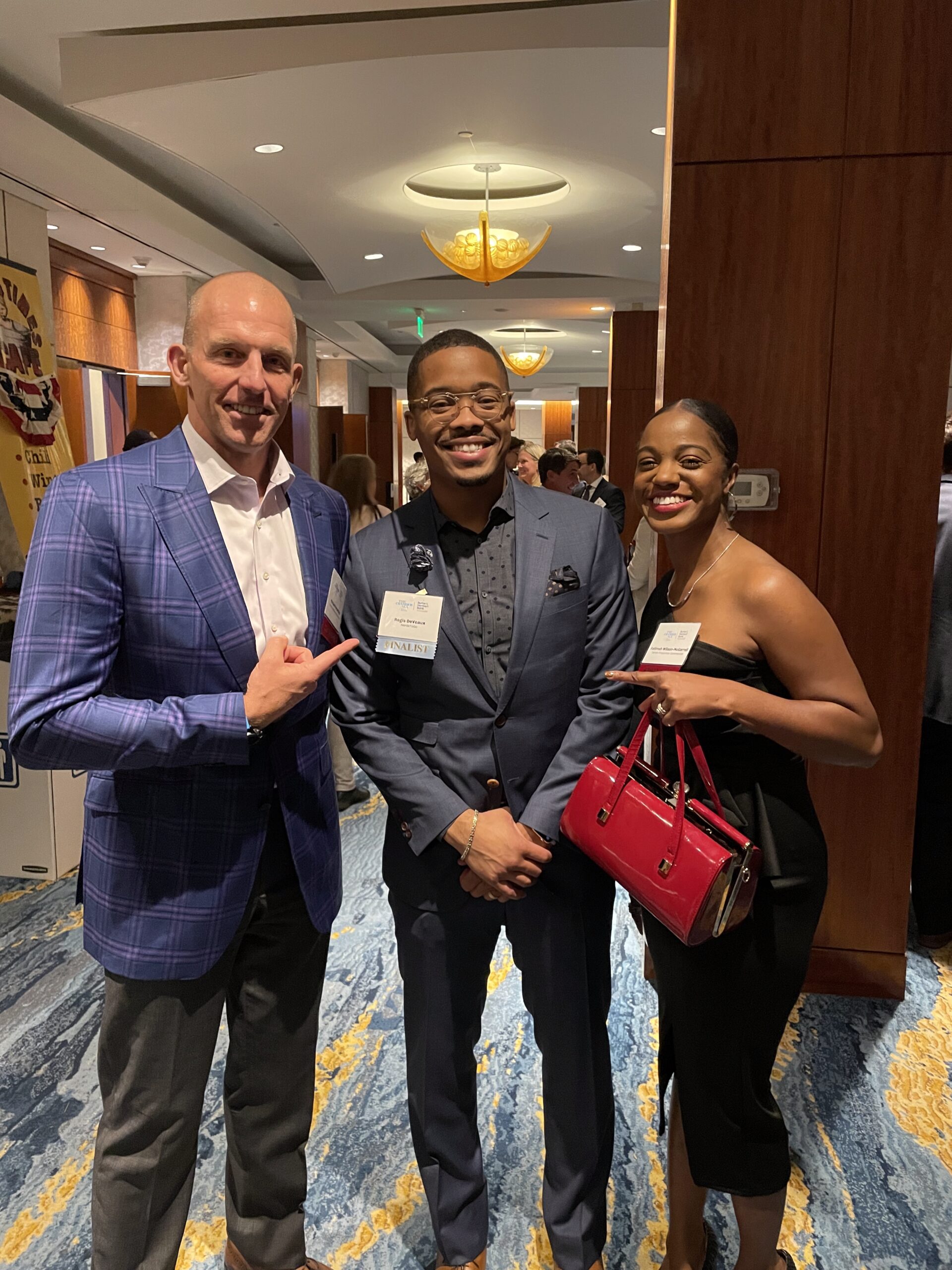 Three people dressed in formal attire smile for a photo at an indoor Tartan Properties event. The man on the left points toward the man in the center, while the woman on the right holds a red purse on a blue and yellow patterned carpet.