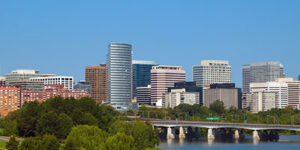 A city skyline with modern high-rise buildings, trees in the foreground, and a bridge crossing over a river under a clear blue sky.