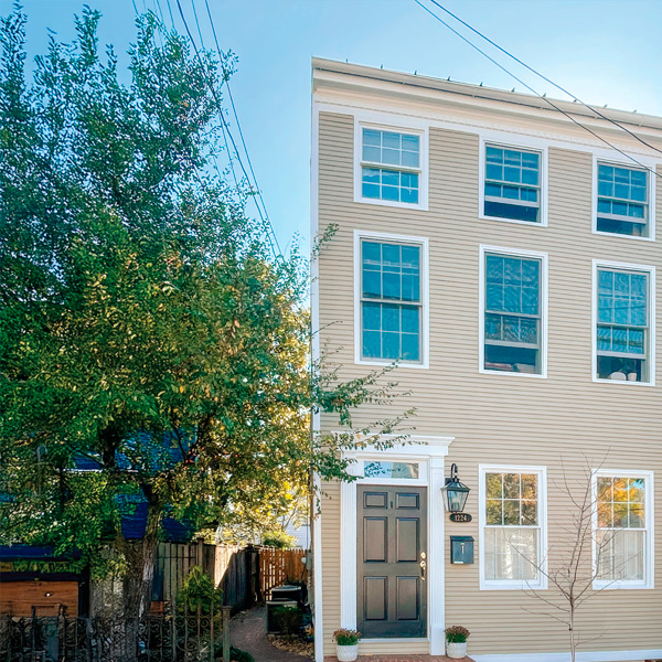A beige three-story townhouse with nine large windows and a black front door. Two potted plants sit by the entrance, and a tree with green leaves stands on the left side, partially shading the house.