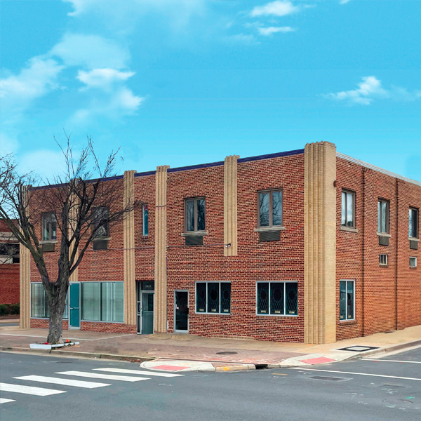 A two-story brick building with large windows on a street corner, featuring vertical tan accents and a leafless tree on the sidewalk, under a bright blue sky with scattered clouds.