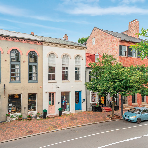 Historic brick buildings with storefronts and arched windows line a quiet street with brick sidewalks. A blue car is parked by the curb, and trees provide shade under a clear sky.