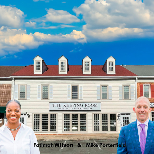 Two people, a woman in a white blazer and a man in a blue suit, stand smiling in front of a large brick building labeled The Keeping Room Fine Home Furnishings under a blue sky with scattered clouds.