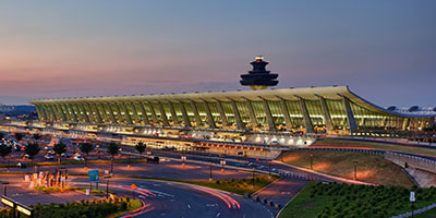 A modern airport terminal with a sweeping, curved roof is illuminated at dusk. A control tower rises above the building, and roads with moving cars and landscaped areas are visible in the foreground.
