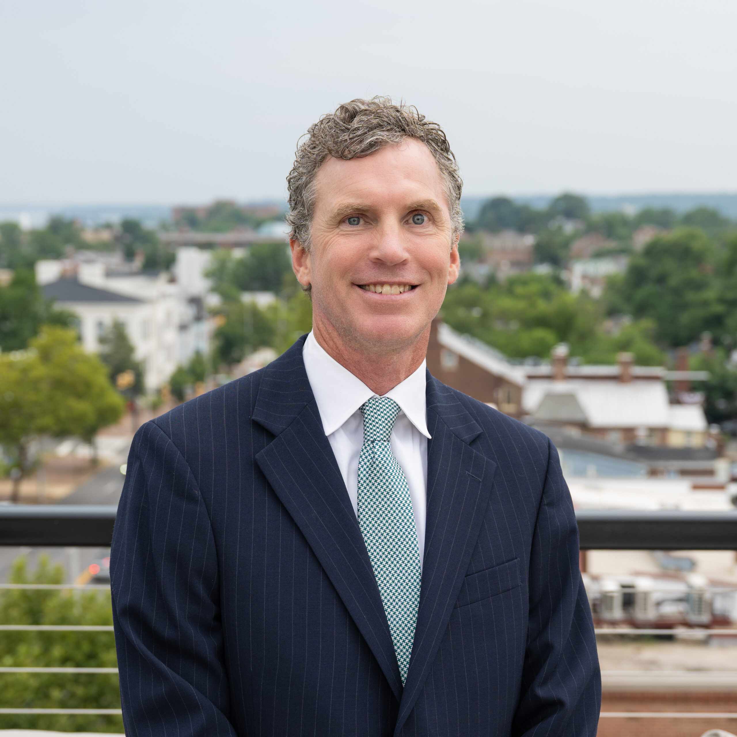 A man in a dark pinstripe suit and light blue tie stands outside, smiling, with a blurred background of trees and buildings.