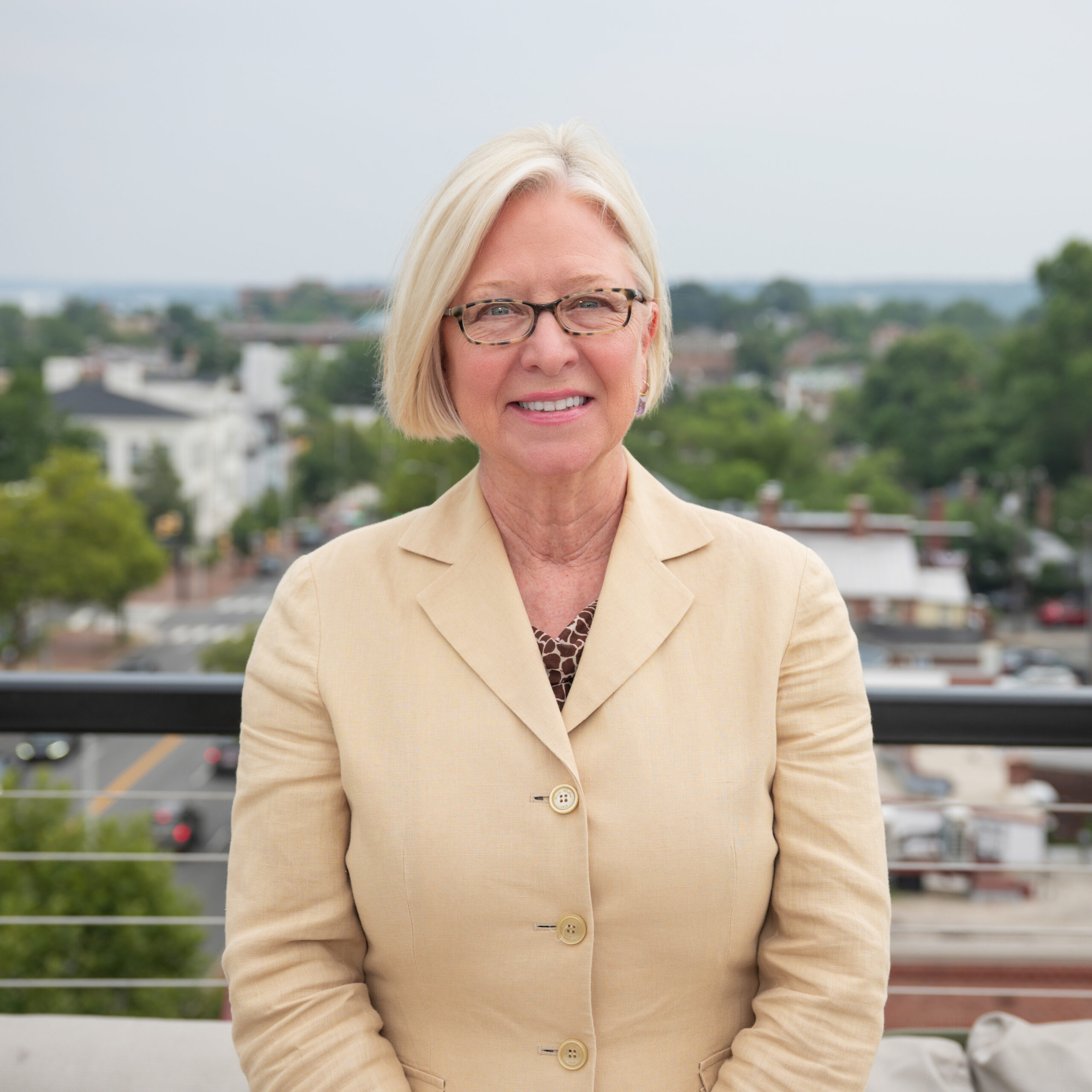 An older woman with short blonde hair and glasses, wearing a beige blazer and patterned blouse, smiles while standing outdoors on a terrace with a blurred cityscape in the background.
