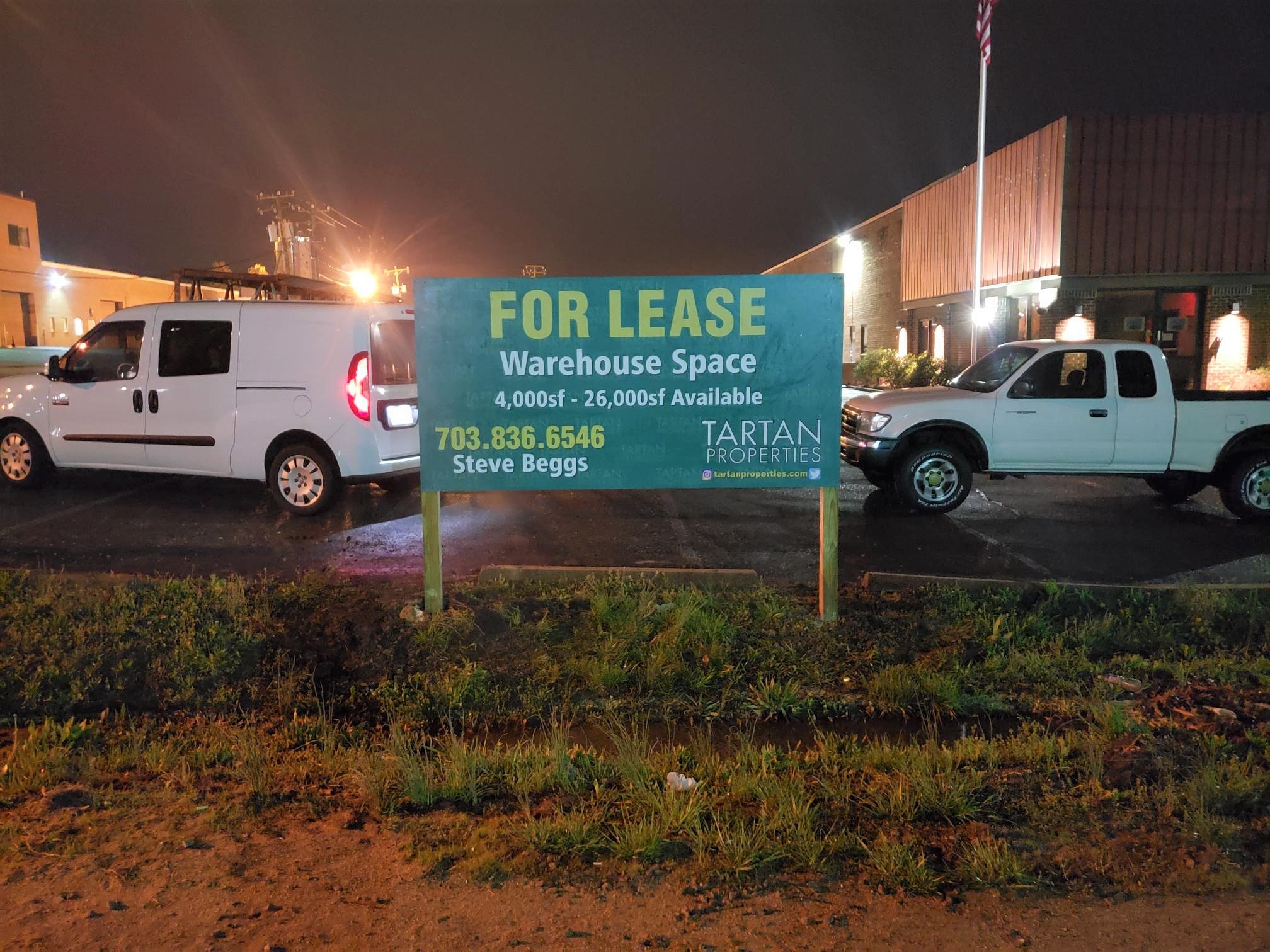A sign reads FOR LEASE Warehouse Space 4,000 SF - 26,000 SF Available. 703.836.6546 Steve Beggs TARTAN PROPERTIES. Two parked vehicles and a building are visible in the background at night.