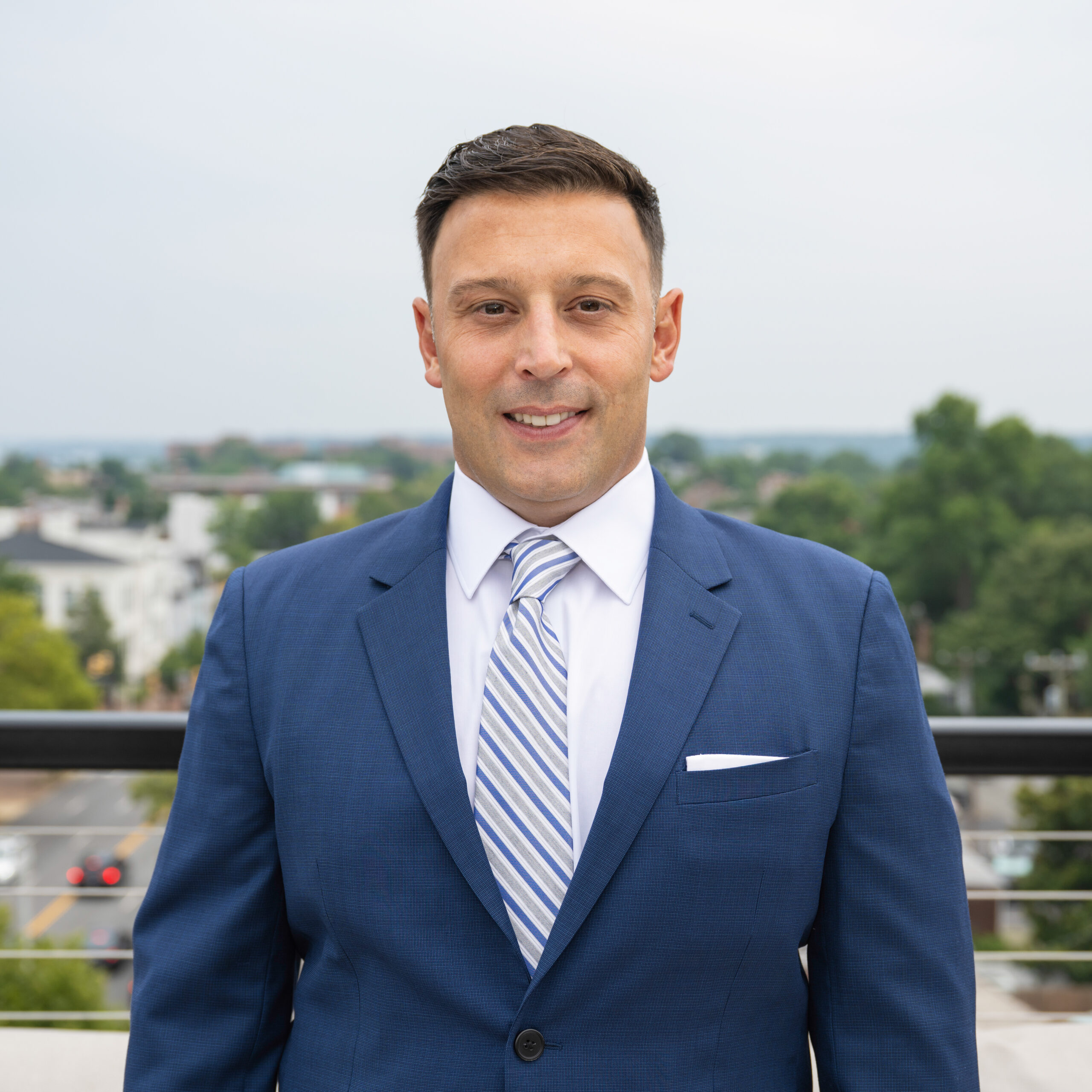 A man in a blue suit and striped tie stands outdoors on a balcony, with a blurred background of trees, buildings, and a street on a cloudy day.