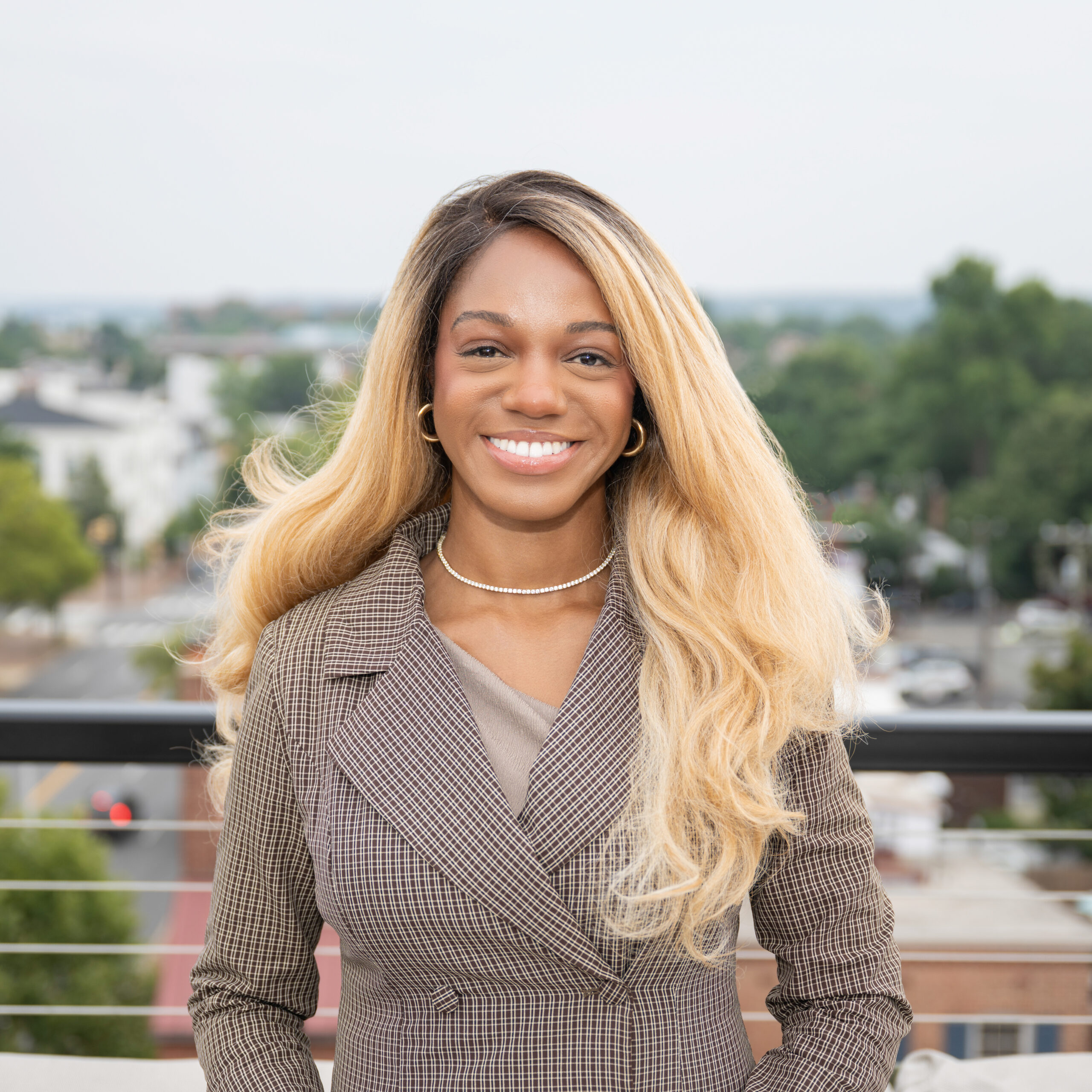 A woman with long, wavy blonde hair smiles while standing outside on a balcony, wearing a brown plaid blazer. The background shows a blurred cityscape with trees and buildings.