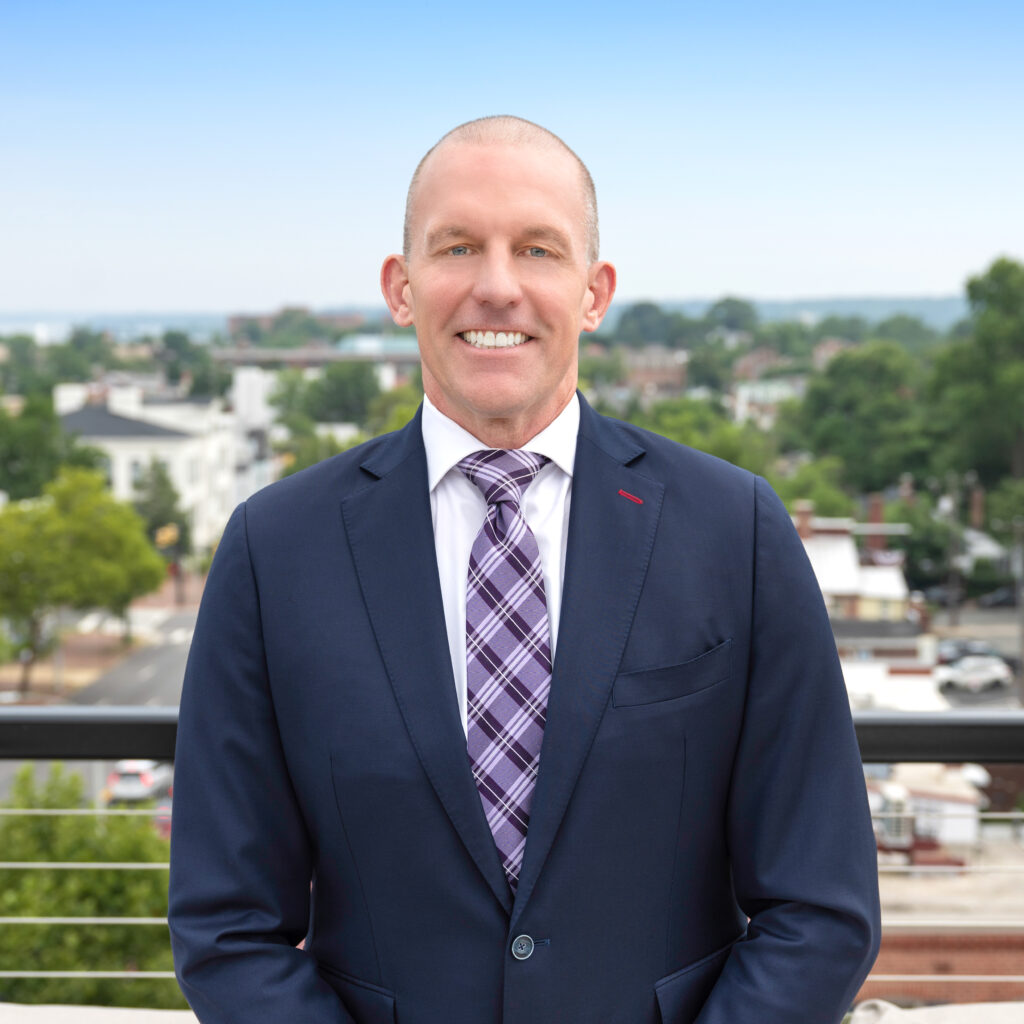 A man in a navy blue suit and plaid tie stands outside on a balcony with a blurred cityscape and greenery in the background under a clear blue sky.