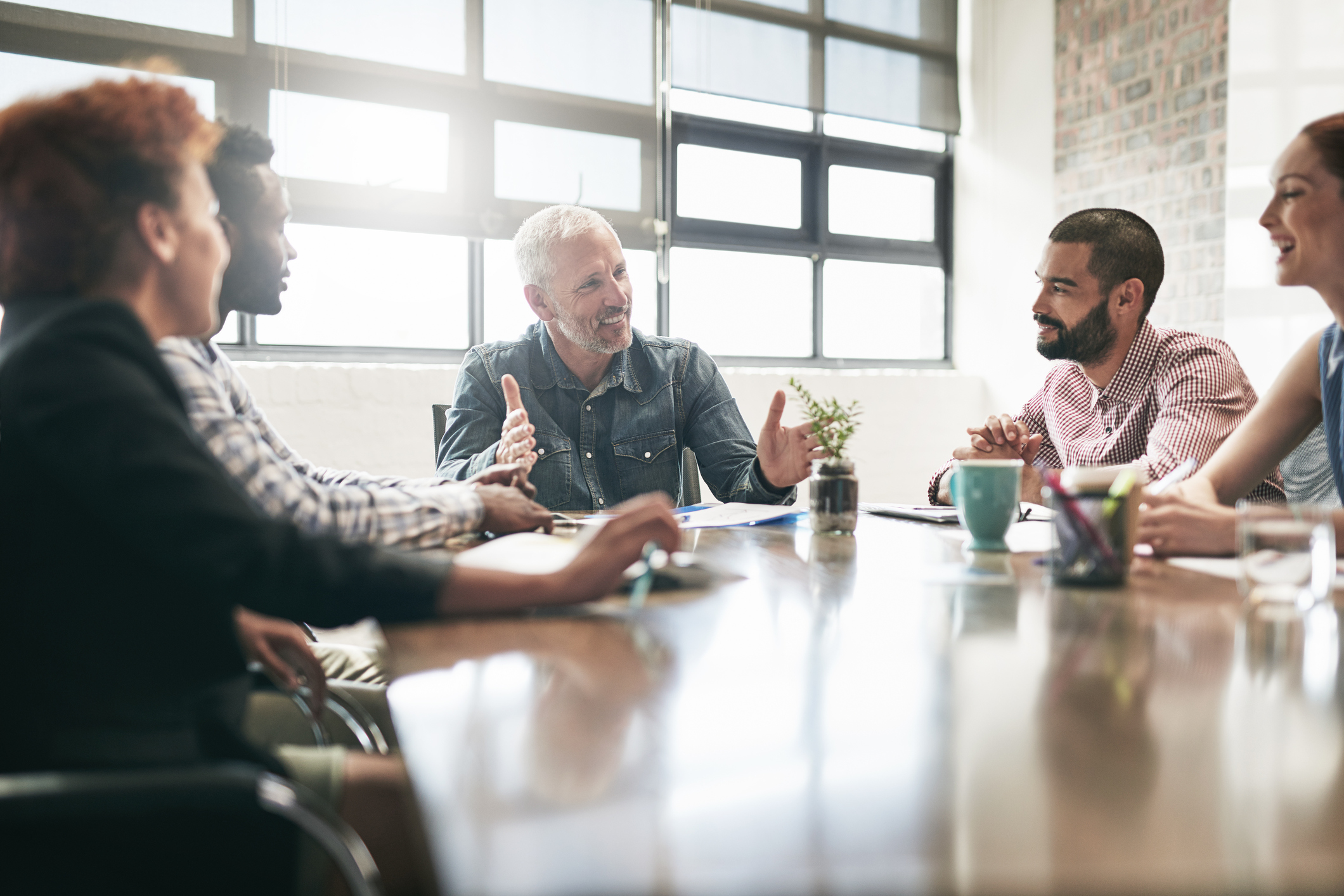 A group of five people sit around a table in a brightly lit office, engaged in a lively discussion. One man in the center gestures while speaking; others listen and smile.
