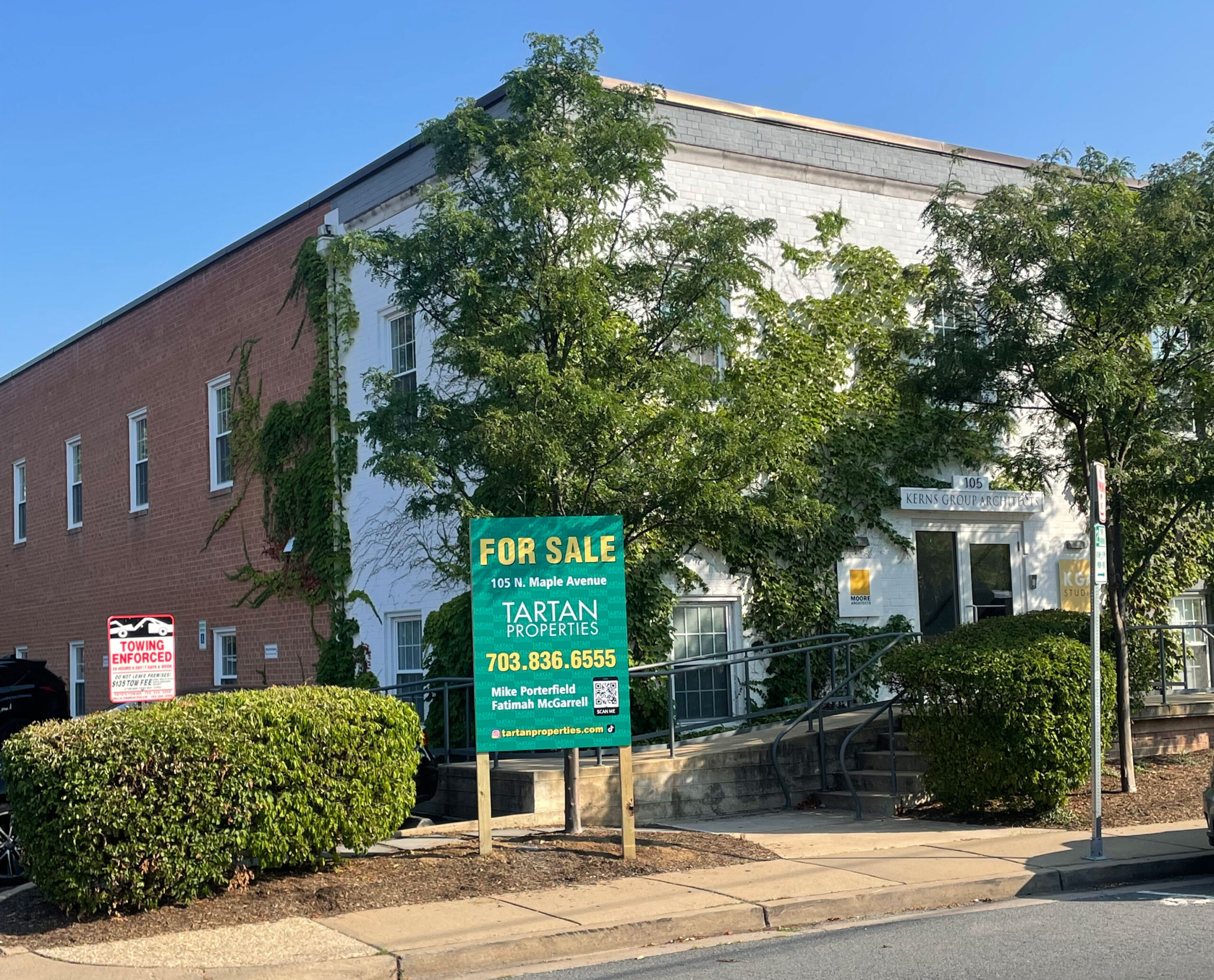 A two-story brick and white building with a large green For Sale sign from Tartan Properties in front, surrounded by bushes and trees, under a clear blue sky.
