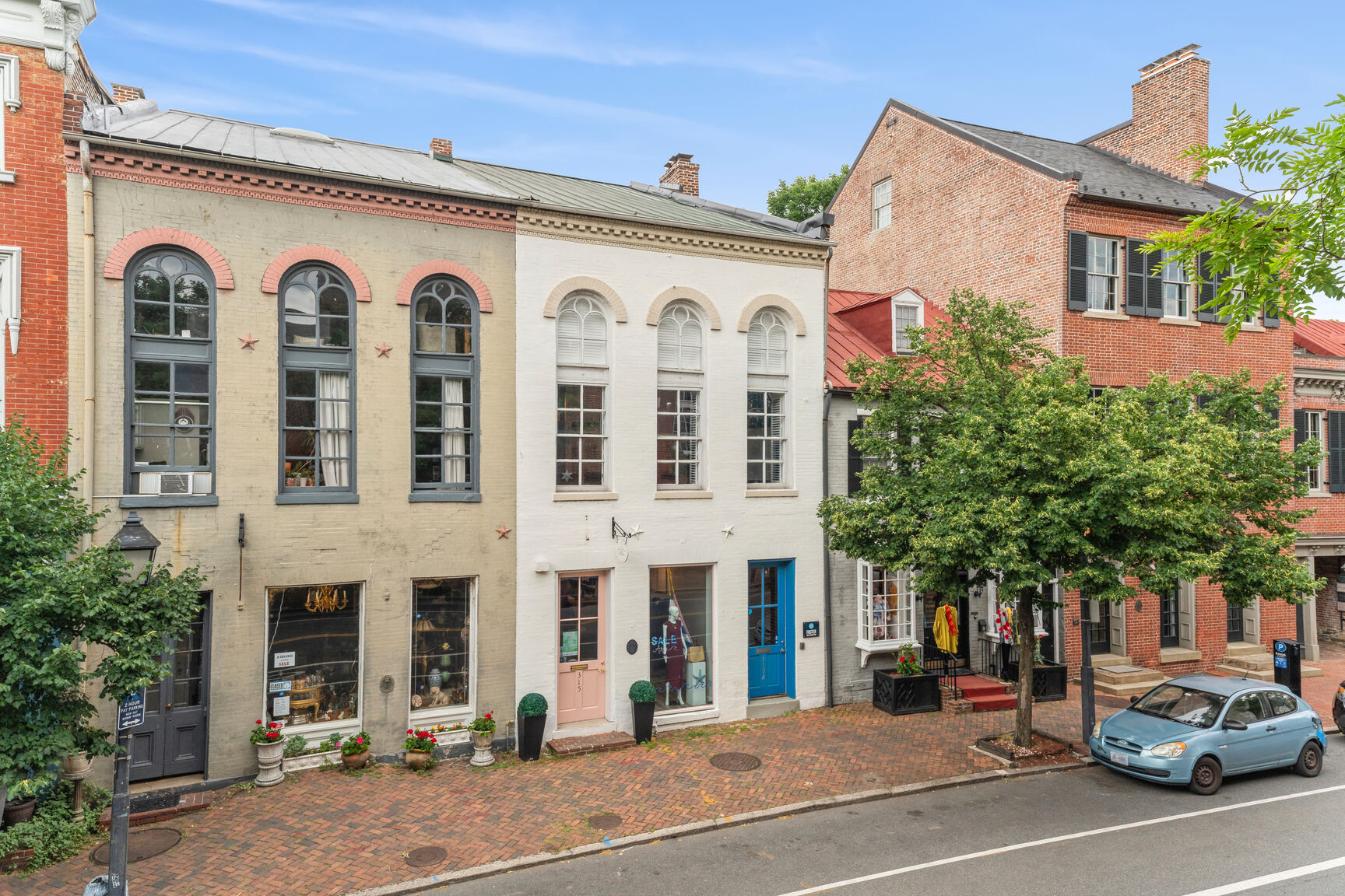 Historic brick buildings with large arched windows line a cobblestone sidewalk on a quiet street; a blue car is parked beside trees, and small shops occupy the ground floors under a clear sky.