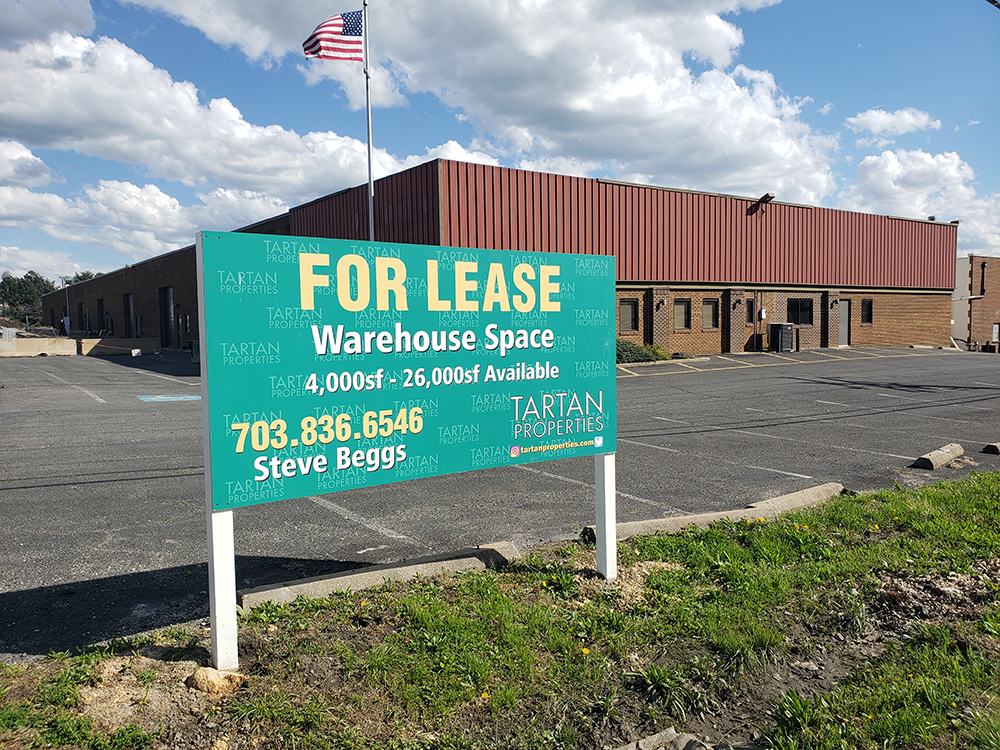 A large green sign in front of a warehouse building reads For Lease, Warehouse Space, 4,000sf - 26,000sf Available with contact information for Tartan Properties and a mostly empty parking lot.