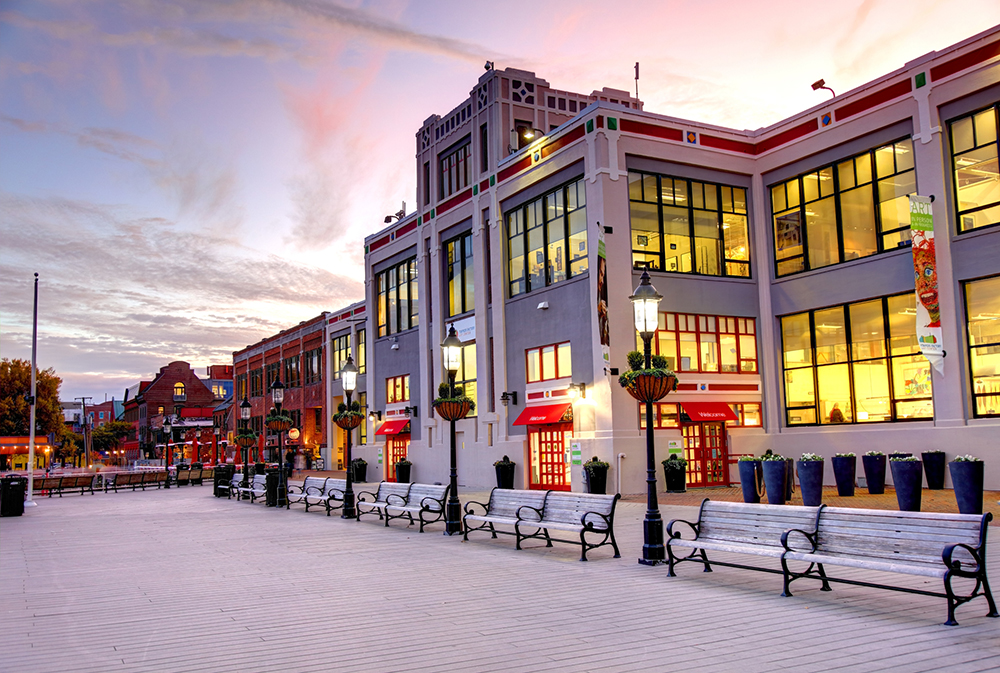 A boardwalk with empty benches lines a waterfront in front of a large, modern building with many windows, glowing warmly at sunset. The sky is pink and purple, and street lamps and planters line the path.