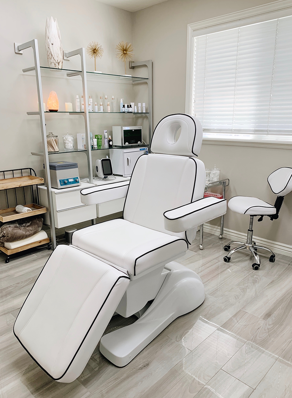 A modern, clean spa or medical treatment room with a white reclining chair, a matching stool, shelves with skincare products, and natural light coming through a window with blinds.