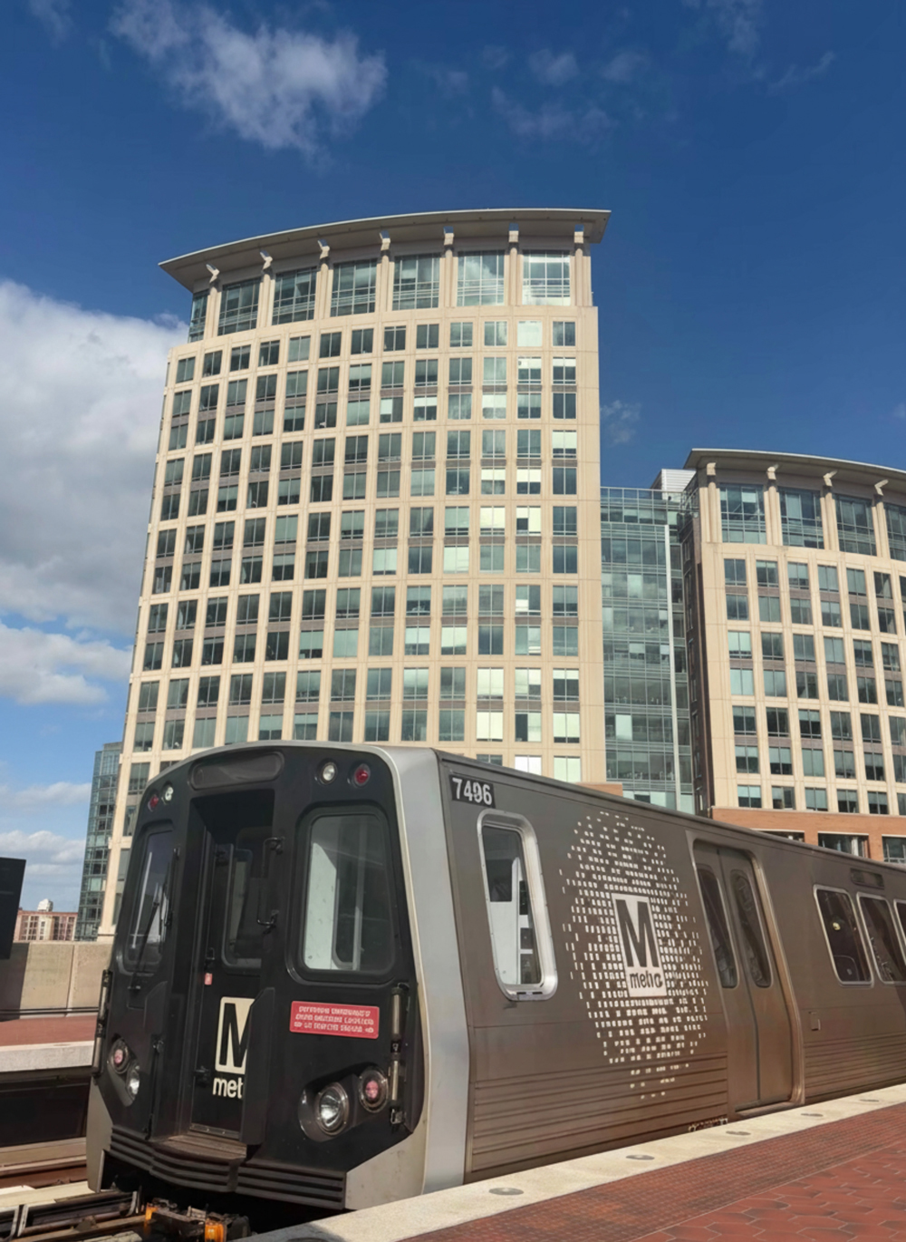 A silver Washington Metro train stopped at an outdoor station platform, with a modern high-rise office building and a blue sky with scattered clouds in the background.