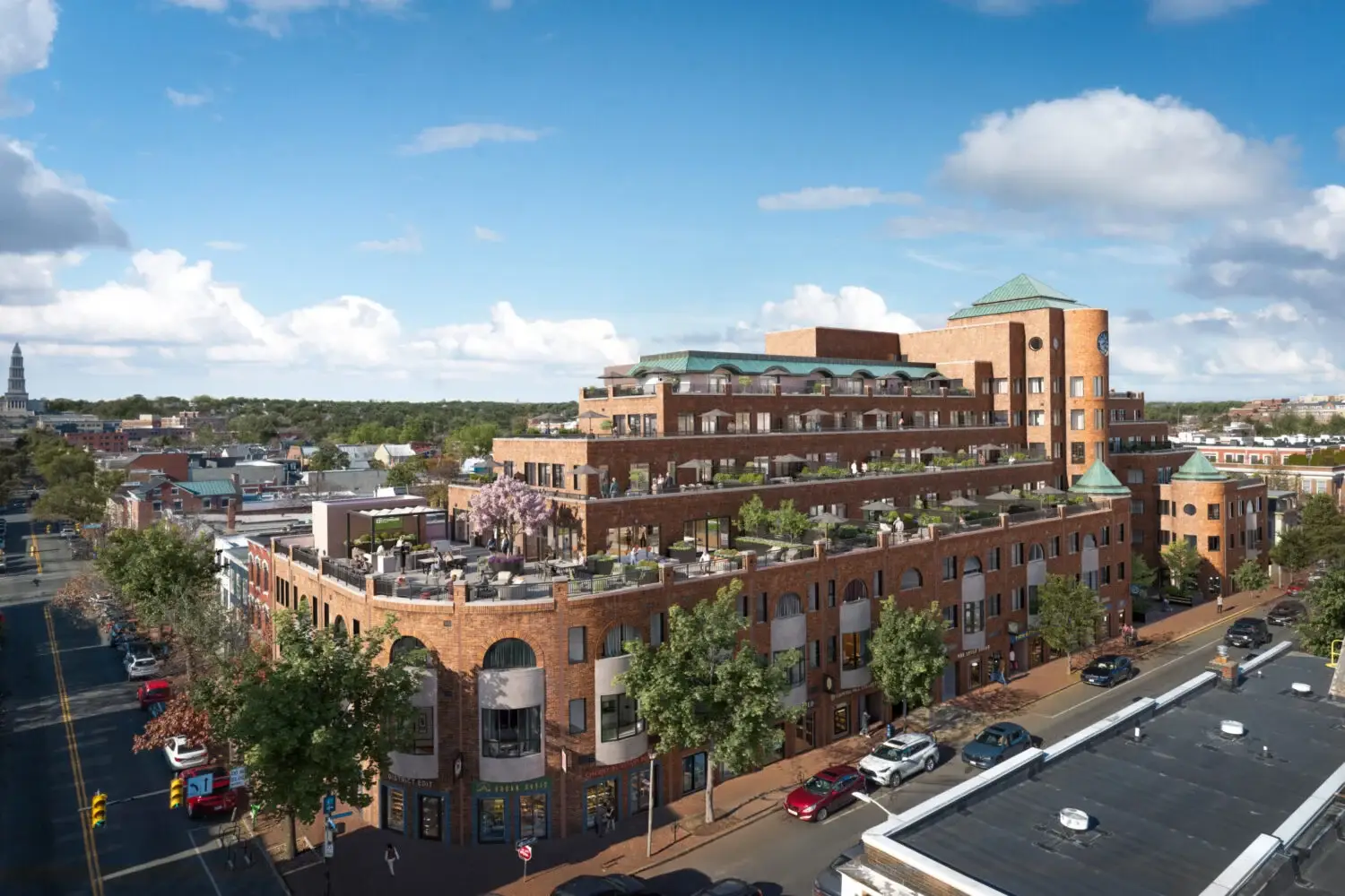 A large, multi-story brick building with multiple terraces and green rooftops stands on a tree-lined street. Cars are parked along the road, and the sky is partly cloudy with city buildings in the background.