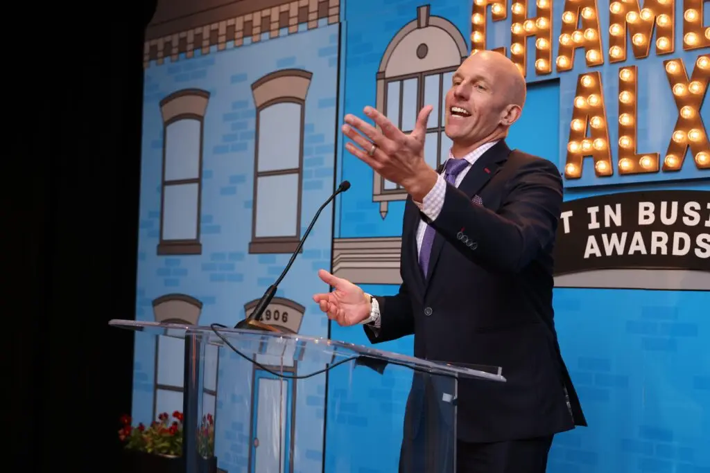 A man in a suit smiles and gestures while speaking at a clear podium on stage, with a blue brick-themed backdrop reading Chamber ALX Best in Business Awards.