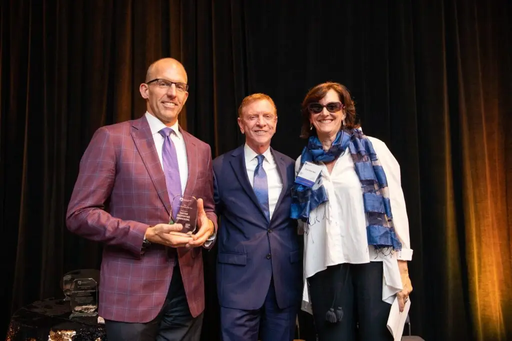 Three people dressed in formal attire stand together and smile at an indoor event; one person on the left holds a glass award. A dark curtain serves as the backdrop.