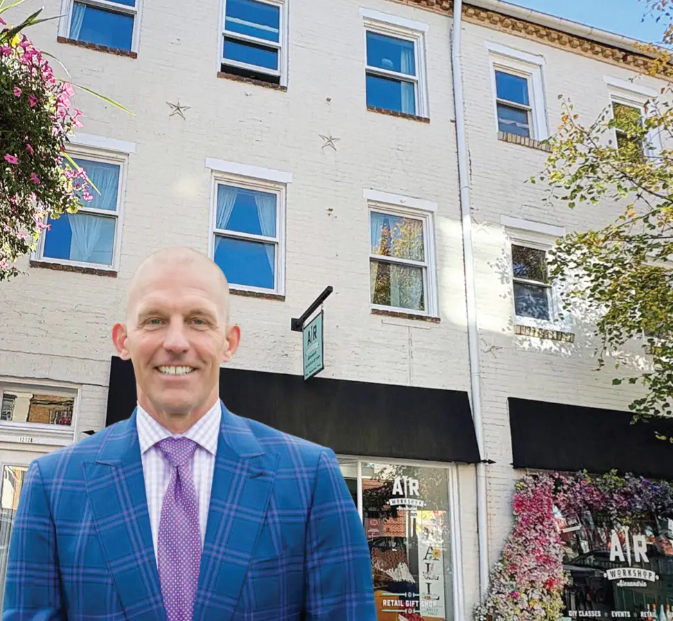 A smiling, bald man in a blue plaid suit stands in front of a white brick building with large windows and a shop called AIR at street level. The scene includes flowers and trees along the sidewalk.