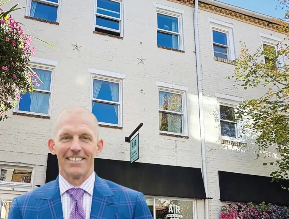 A man in a blue plaid suit and pink tie stands smiling in front of a three-story white brick building with large storefront windows and flower decorations.
