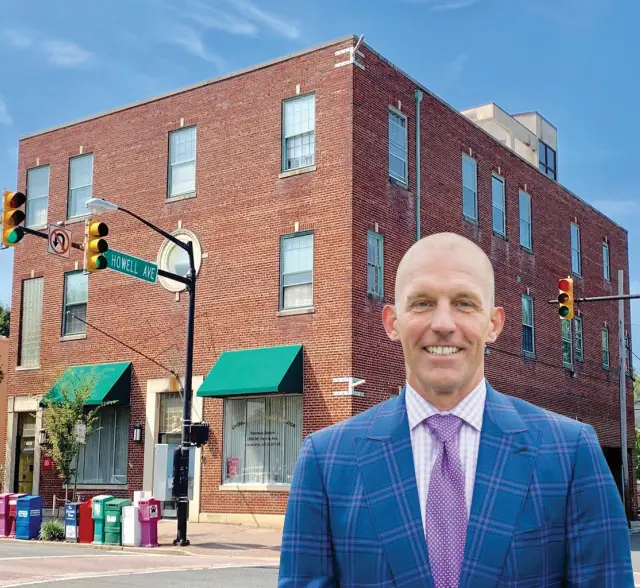 A smiling man in a blue plaid suit stands at a street corner with a brick building, green awnings, traffic lights, and newspaper boxes in the background. The street signs read Howell Ave. and Mt. Vernon Ave.