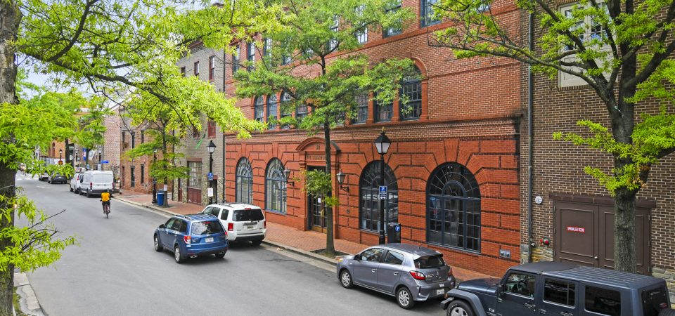 A red brick building with large arched windows lines a tree-shaded street where cars are parked and a cyclist rides past on the left. The scene is calm and urban, with a mix of sunlight and shade.