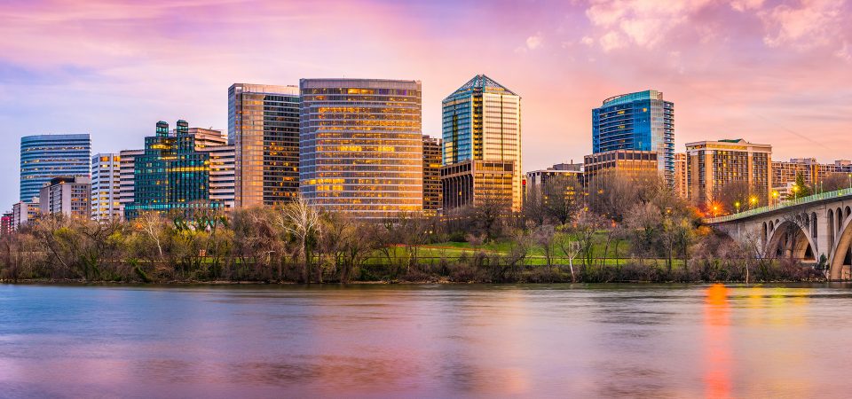 A city skyline with modern buildings, green trees, and a bridge, reflected in a calm river at sunset with a colorful pink and purple sky.
